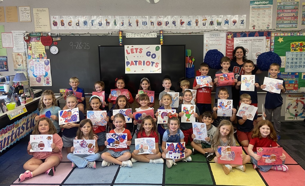 A class of first grade students pose in a group with their teacher smiling at the camera, the students are holding football and cheer leading signs in their classroom on the rug with a sign that says Let’s go patriots behind them.