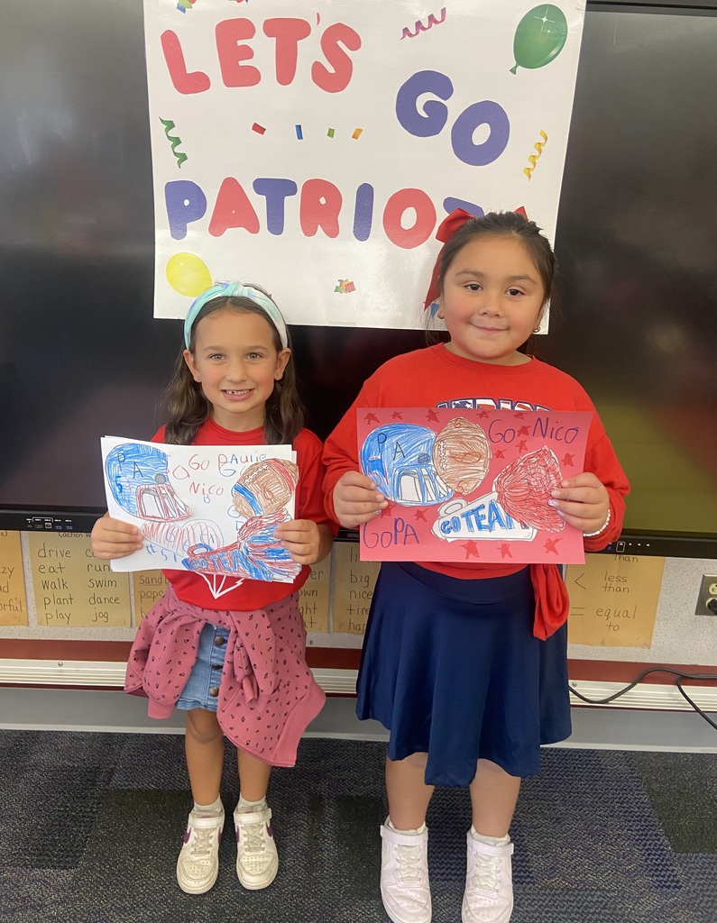 Two girls hold red, white and blue handmade signs that say go team. In front of a sign that says Lets go Patriots. 