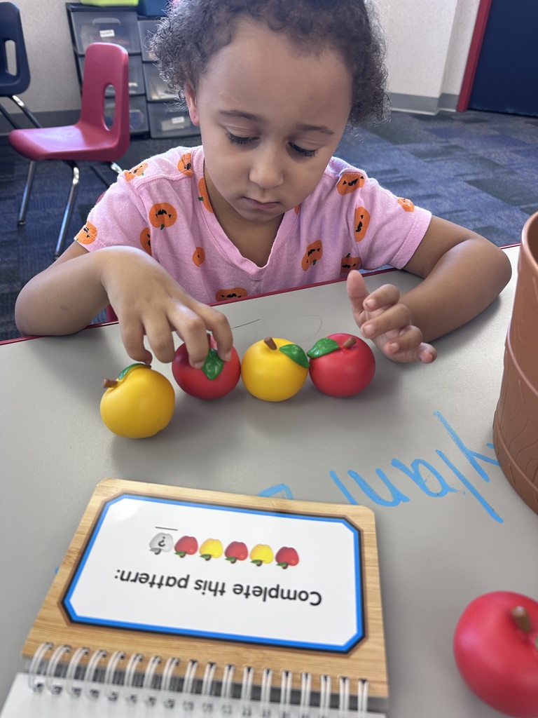 A girl at a classroom table ordering math manipulatives with a book that says complete the pattern.