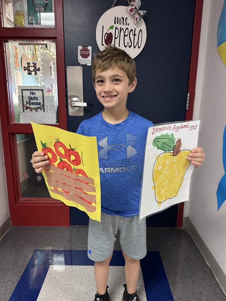 A boy smiling holding a painting if apples in a basket and an apple coloring sheet colored with yellow, brown and green dot markers.