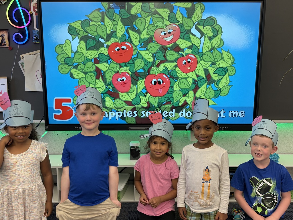 Four students wearing paper johnny appleseed hats in front of a smartboard with 5 apple song on the smart board behind them.