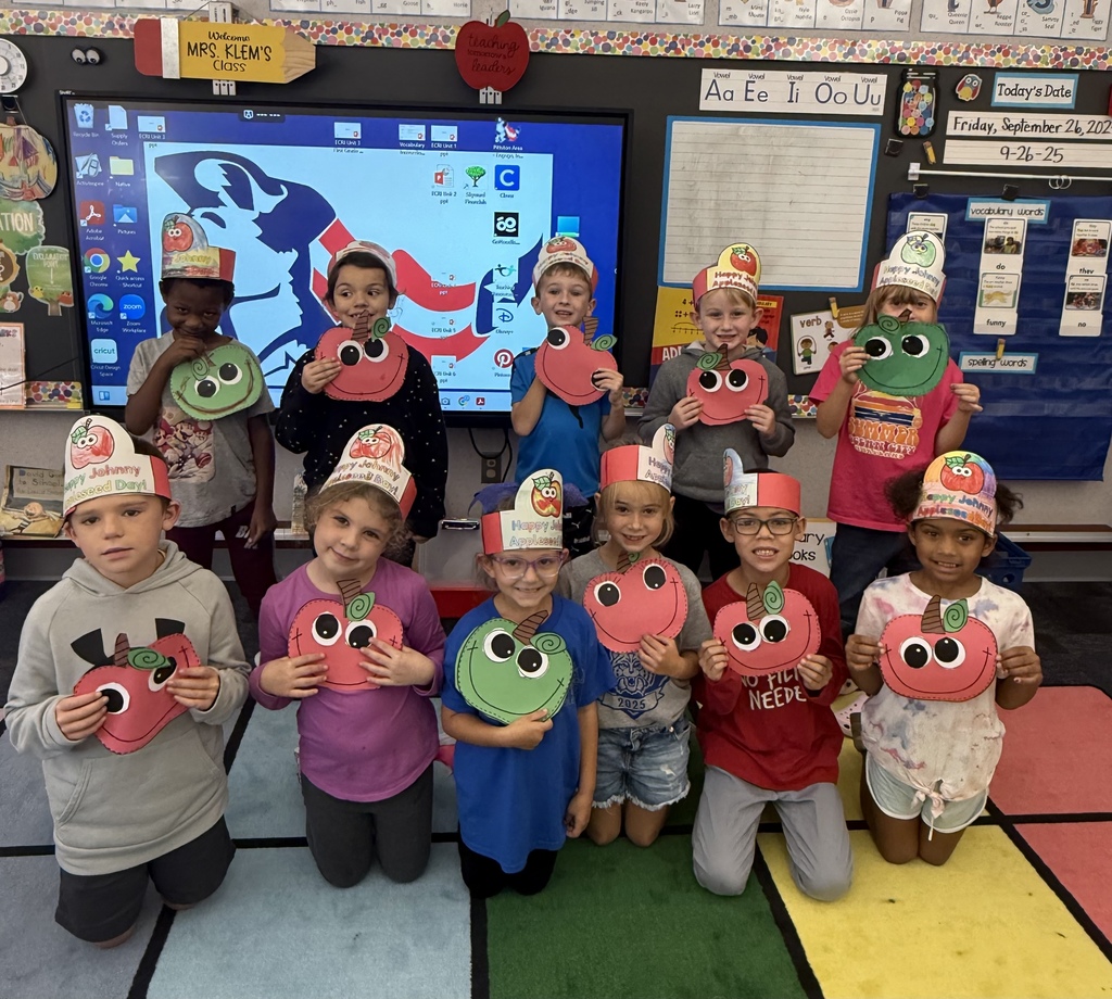 Students posing for a group picture holding  red and green apple crafts with eyes and wearing apple crowns.