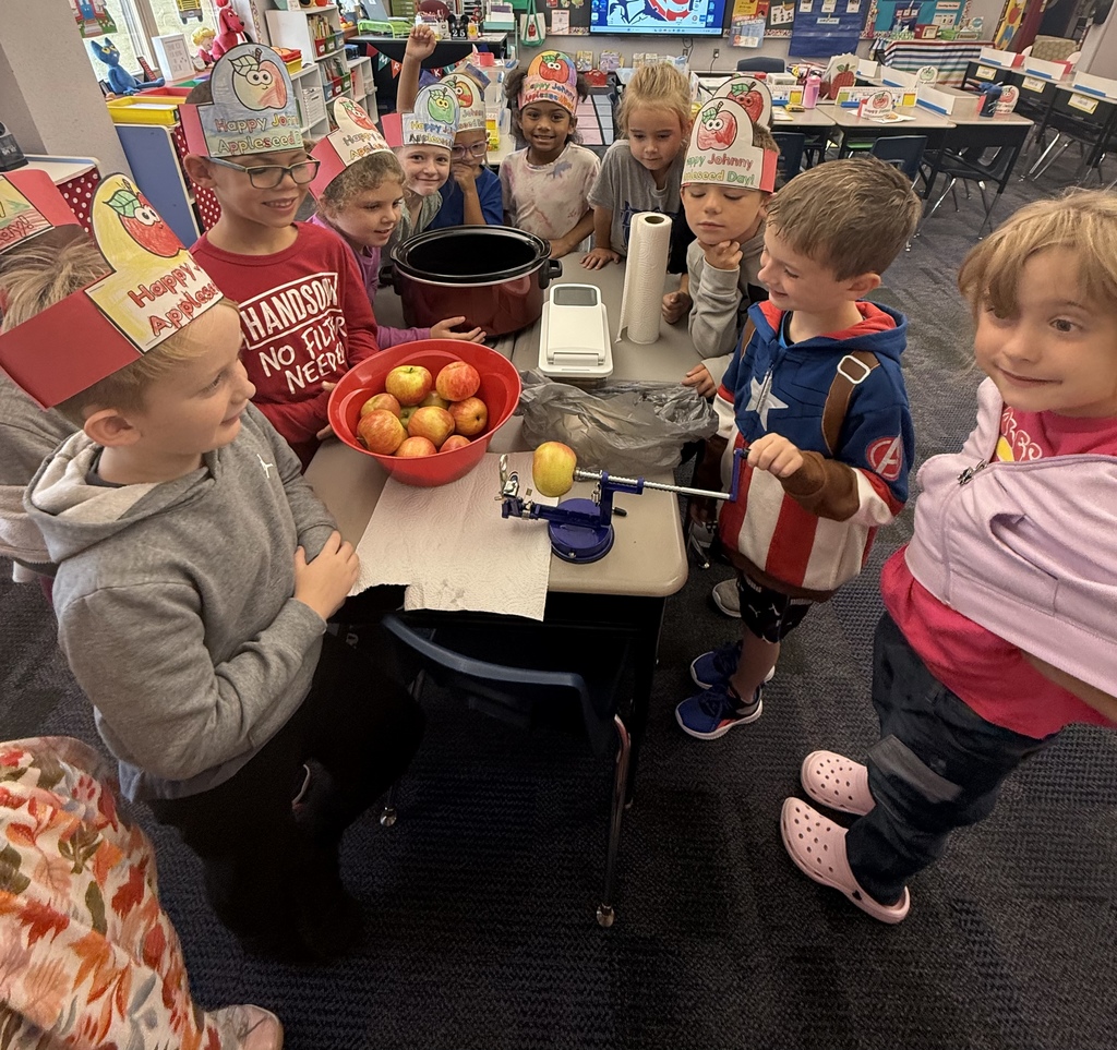 Studnets standing around a table with a red bowl of apples, one student is peeling an apple with an apple peeler. 