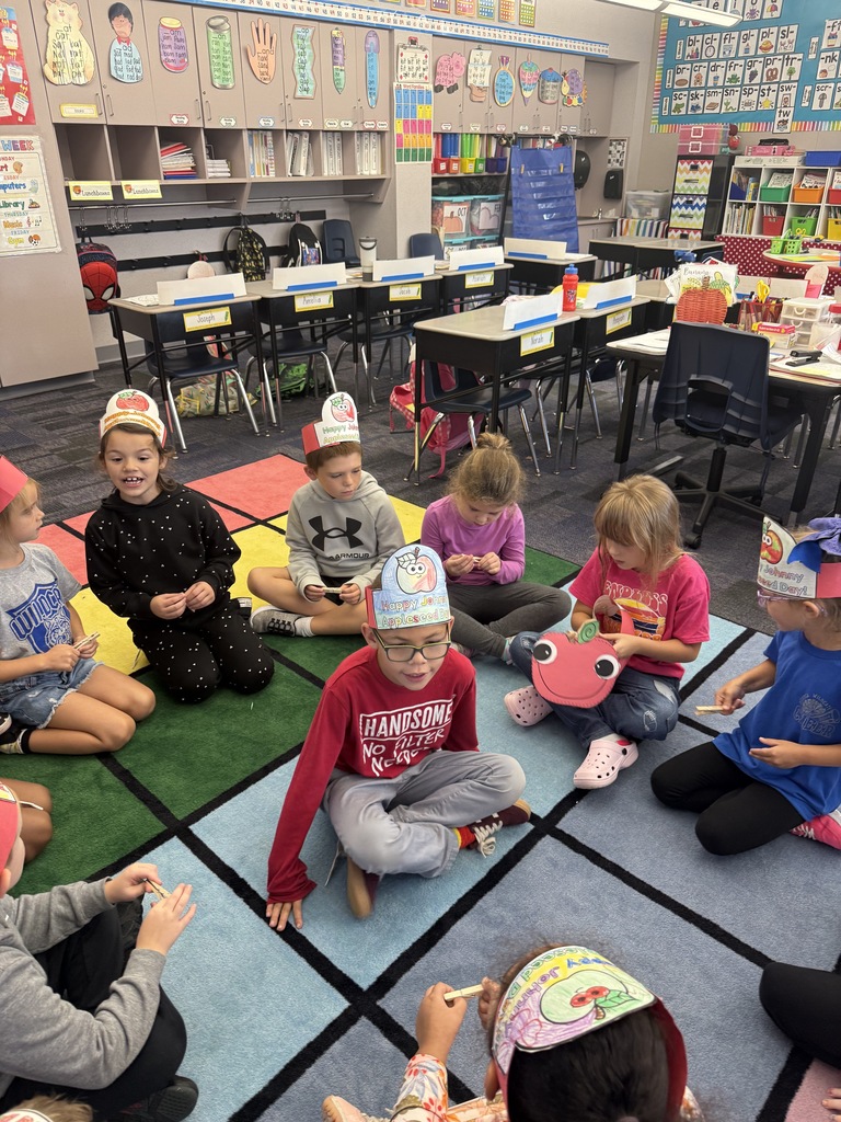 Students sitting in a circle with one student sitting in the middle. One  student is holding a paper apple craft. 