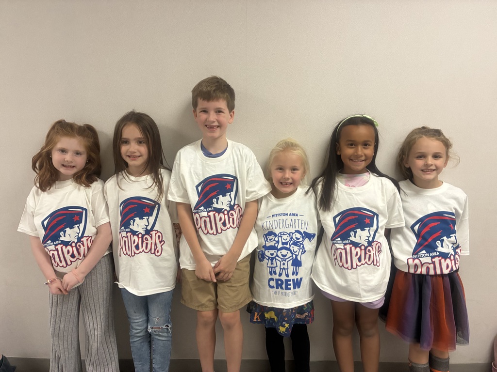 A group of students smiing at the camera in a school hallway in white t shirts with red and blue patriot logos. 