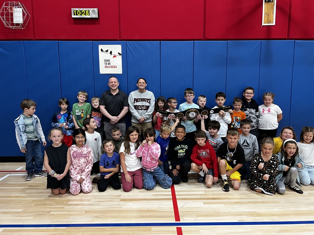 Mrs. Melvin’s 2nd grade class posing for a picture with serious wrestling faces. Two wrestling coaches smile in the picture and the kids are holding a wrestling belt. 