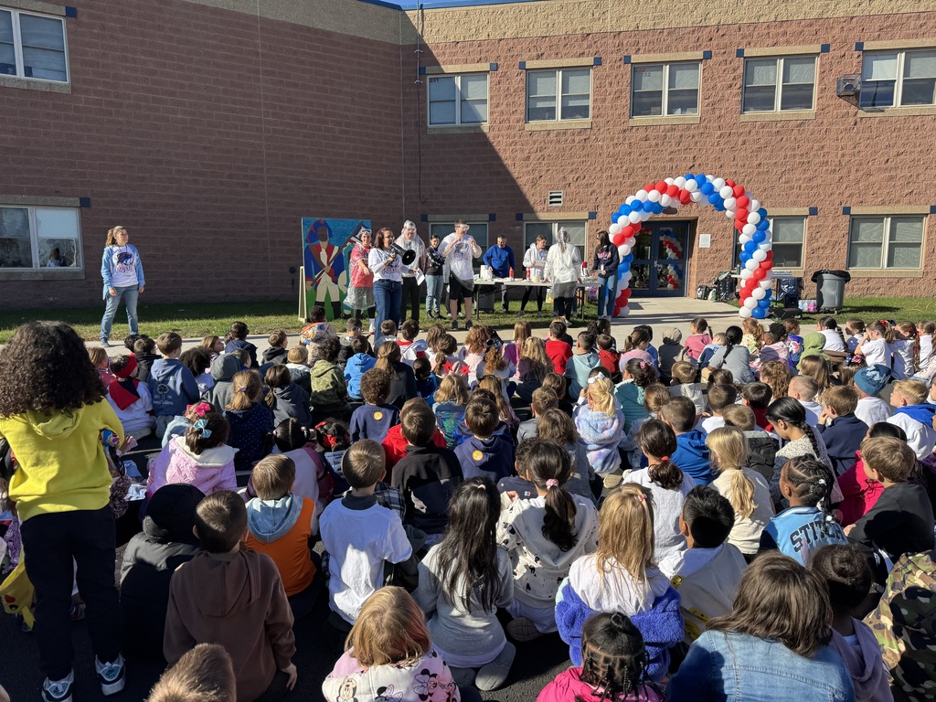 Group of students seated looking at teachers in front of red, white and blue balloon arch.