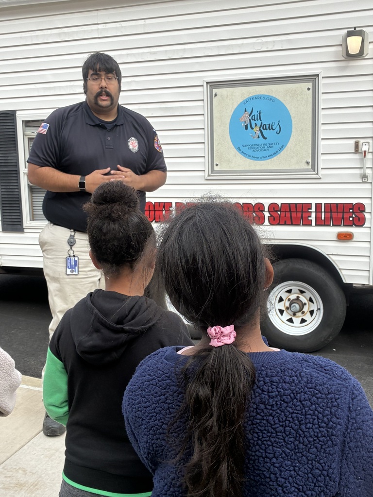 A fireman talking to a group of students in-front of a smokehouse.