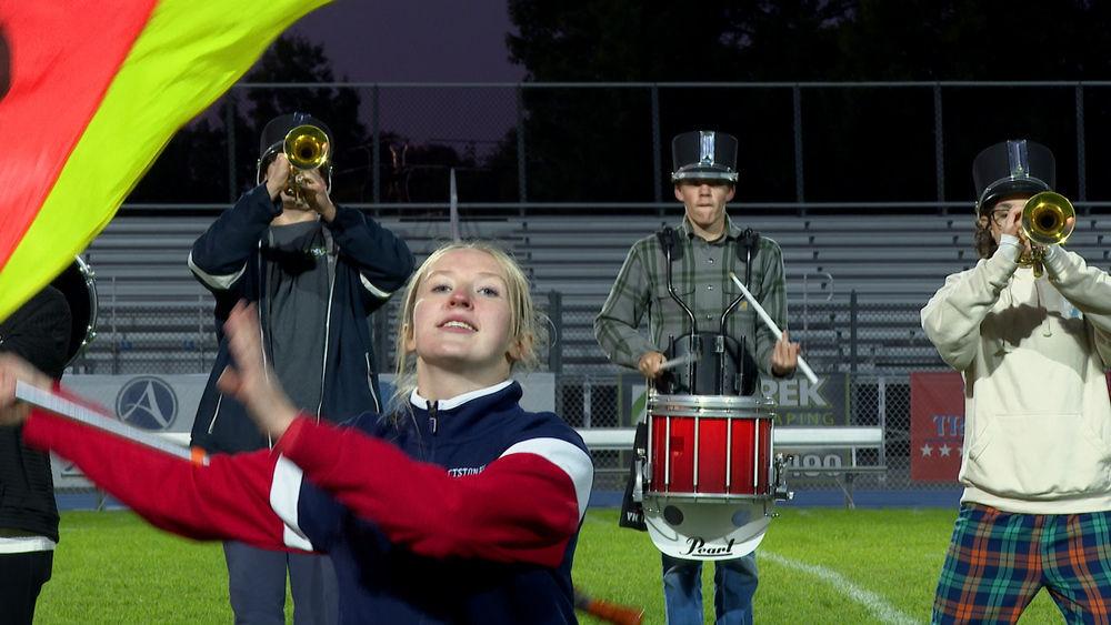 A marching band on field a girl with a flag, a boy with a trumpet and a boy with drums