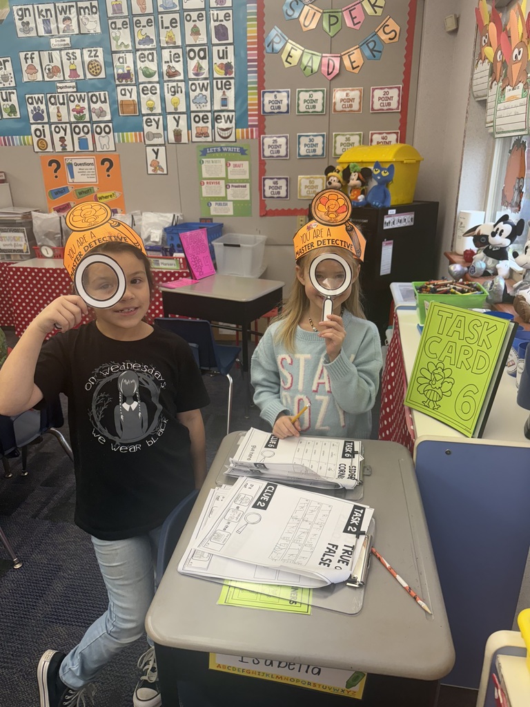 First grade students in a classroom wearing orange paper turkey detective headbands and holding white magnifying glasses.