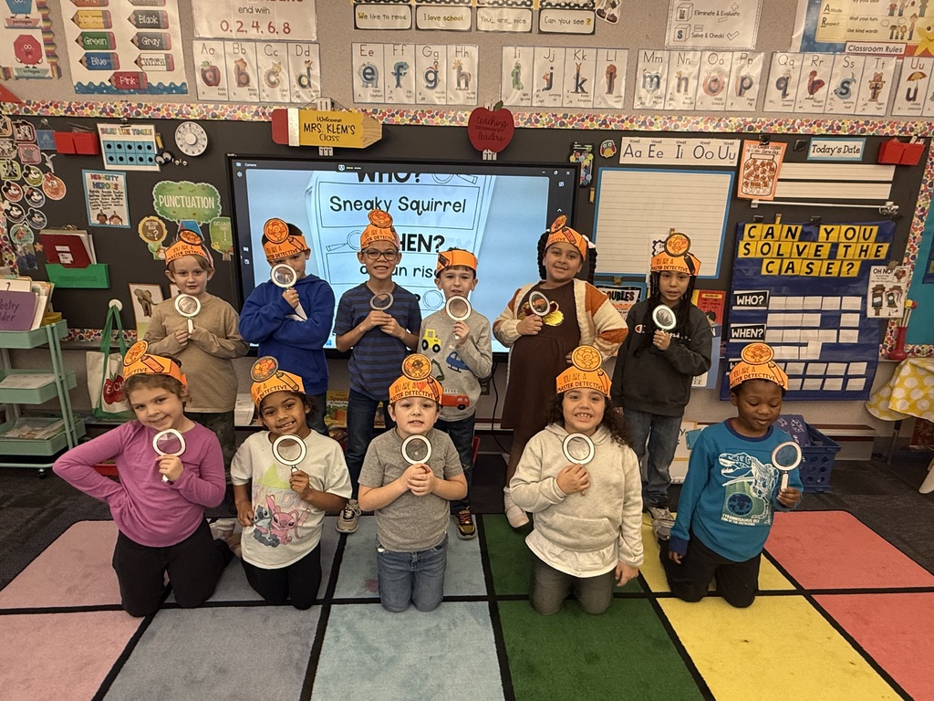 First grade students in a classroom wearing orange paper turkey detective headbands and holding white magnifying glasses.