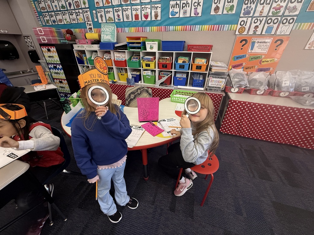 First grade students in a classroom wearing orange paper turkey detective headbands and holding white magnifying glasses.