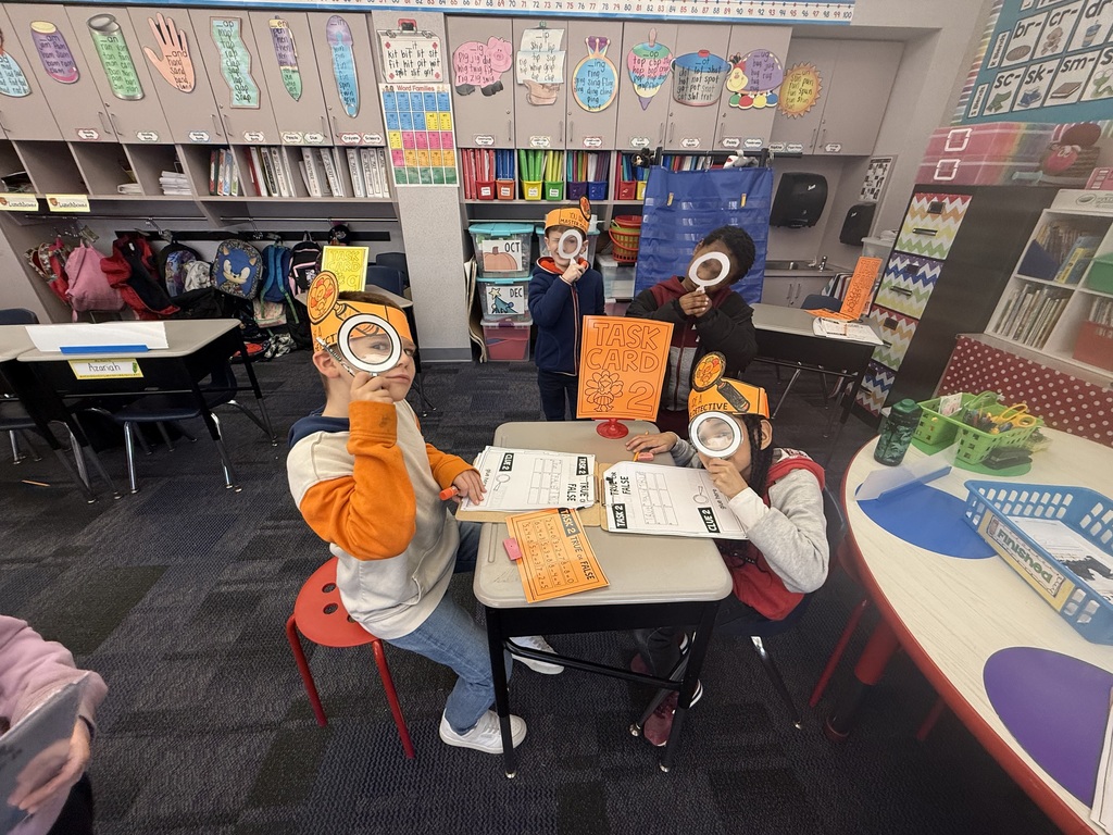 First grade students in a classroom wearing orange paper turkey detective headbands and holding white magnifying glasses.