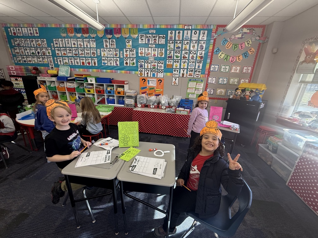First grade students in a classroom wearing orange paper turkey detective headbands and holding white magnifying glasses.