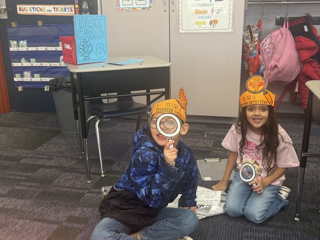 First grade students in a classroom wearing orange paper turkey detective headbands and holding white magnifying glasses.