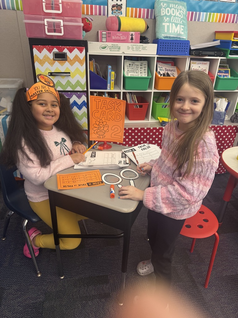 First grade students in a classroom wearing orange paper turkey detective headbands and holding white magnifying glasses.