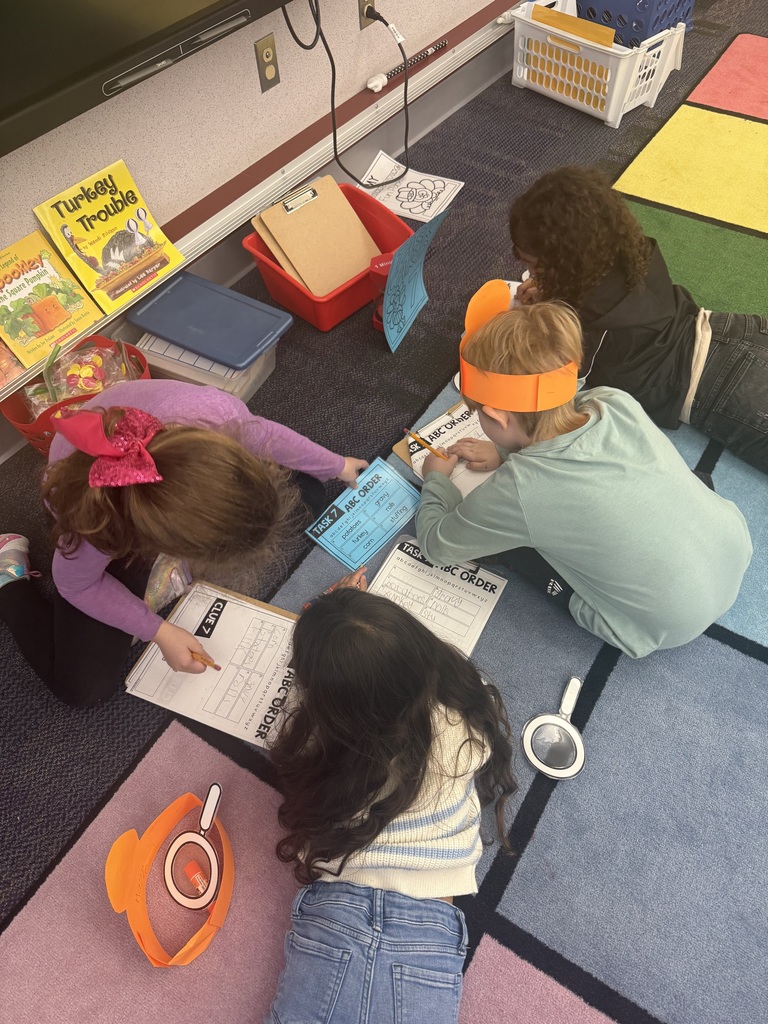 First grade students in a classroom wearing orange paper turkey detective headbands and holding white magnifying glasses.