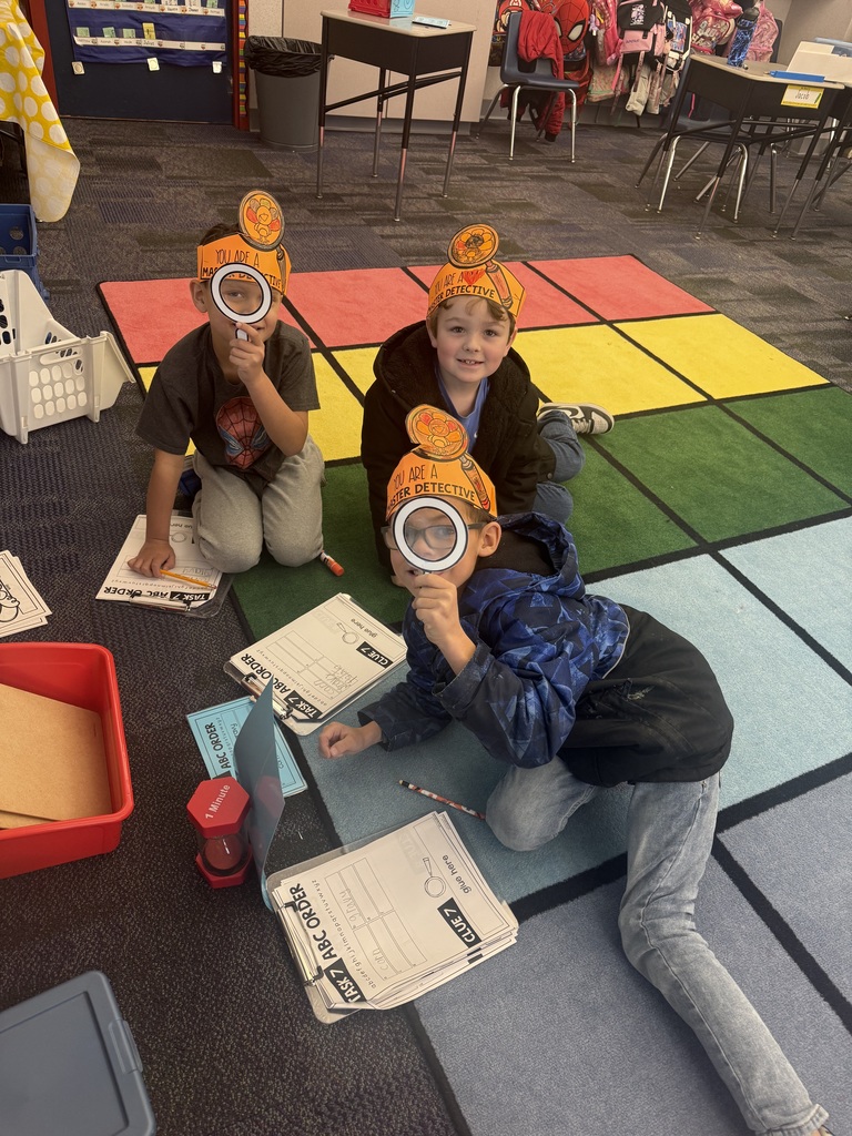 First grade students in a classroom wearing orange paper turkey detective headbands and holding white magnifying glasses.