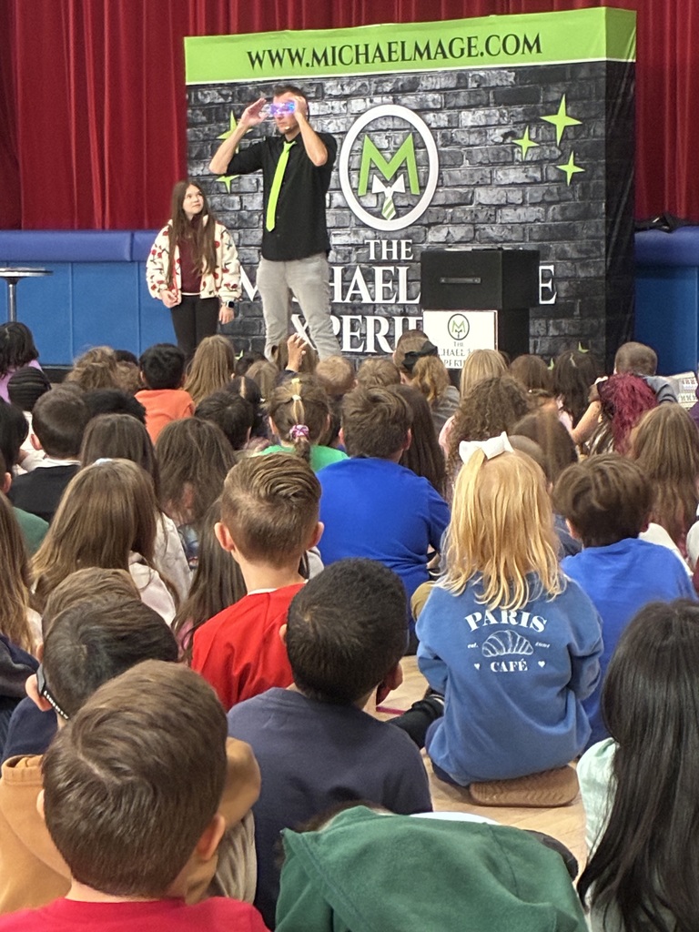 Kids seated in a school gym at a magic show in front of a red and blue stage.