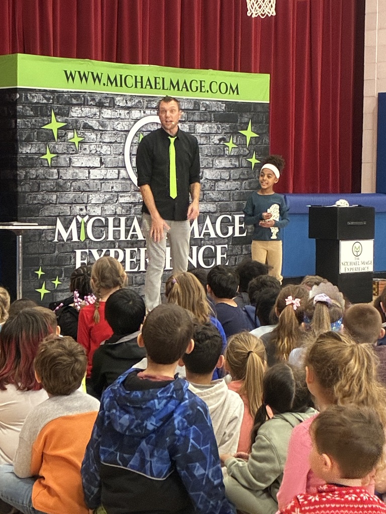 Kids seated in a school gym at a magic show in front of a red and blue stage.