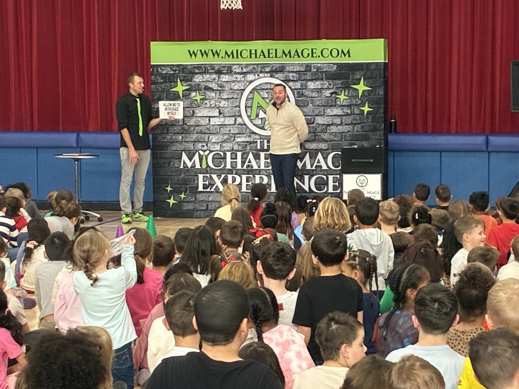 Kids seated in a school gym at a magic show in front of a red and blue stage.