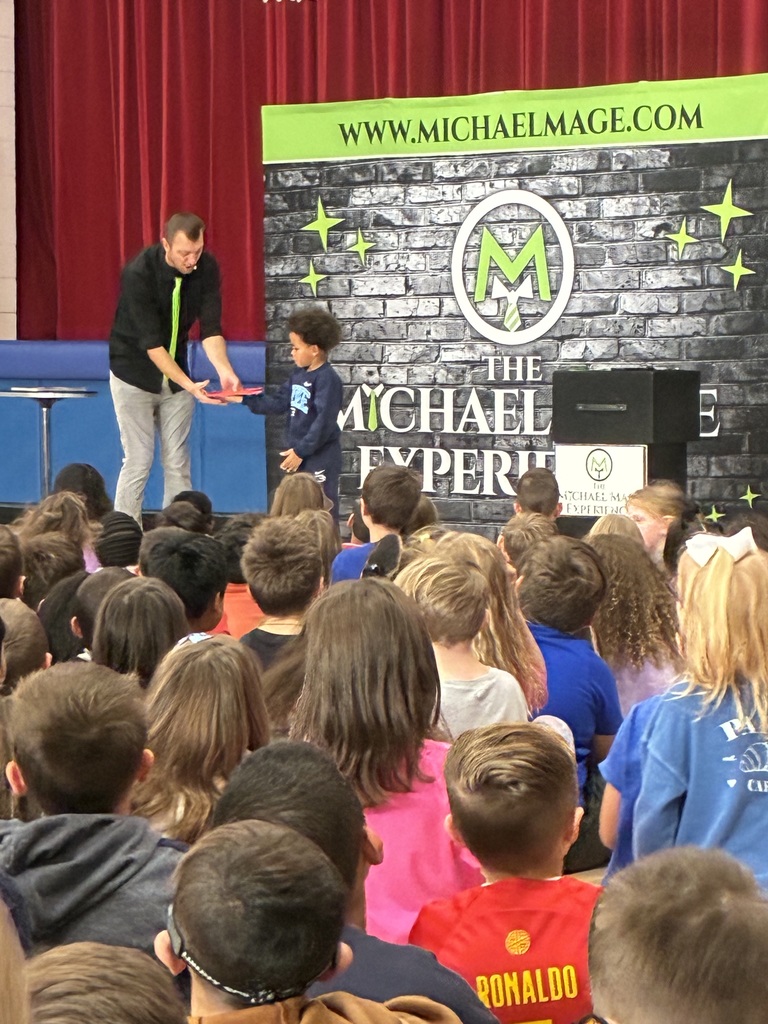 Kids seated in a school gym at a magic show in front of a red and blue stage.