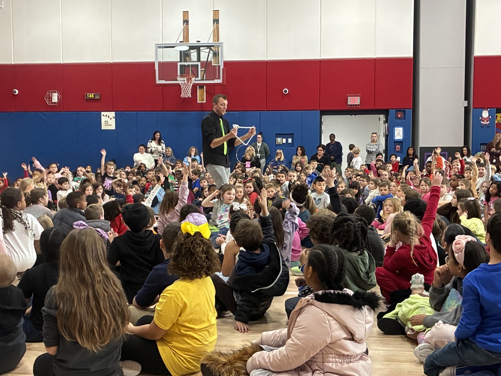 Kids seated in a school gym at a magic show in front of a red and blue stage.