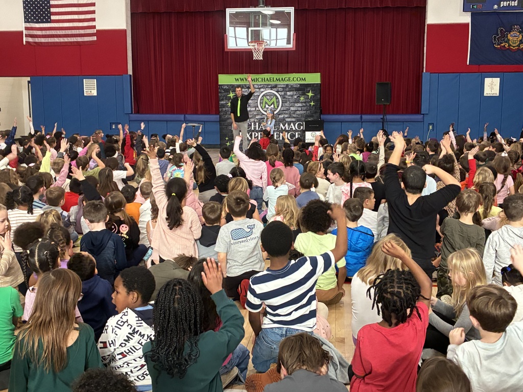 Kids seated in a school gym at a magic show in front of a red and blue stage.