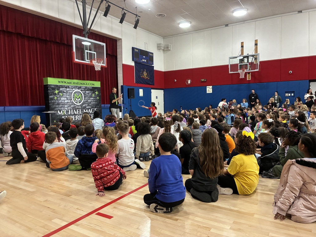 Kids seated in a school gym at a magic show in front of a red and blue stage.