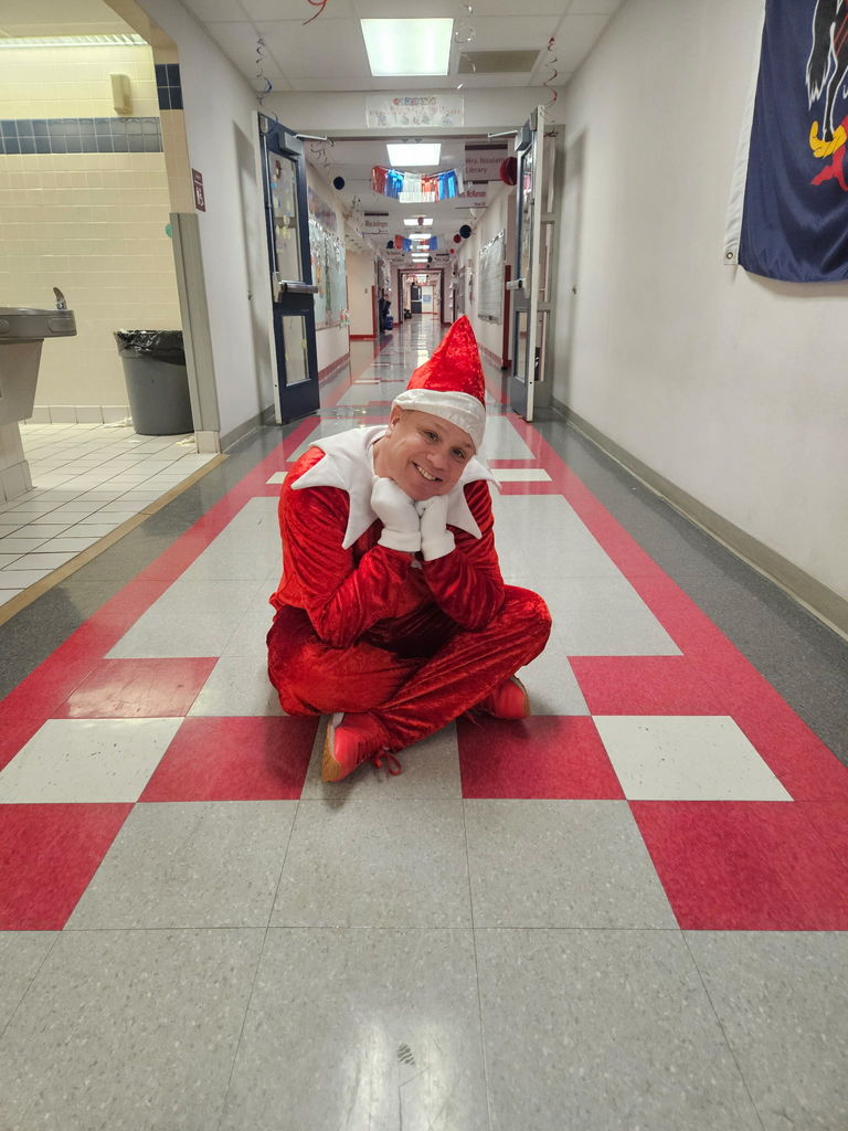 A man dressed as an elf on the shelf seated in a school hallway. 