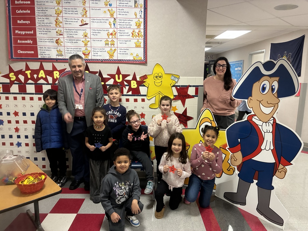 A group of elementary students posing in front of star club bingo board with Mr. savokinas and Mrs. Ferentino 