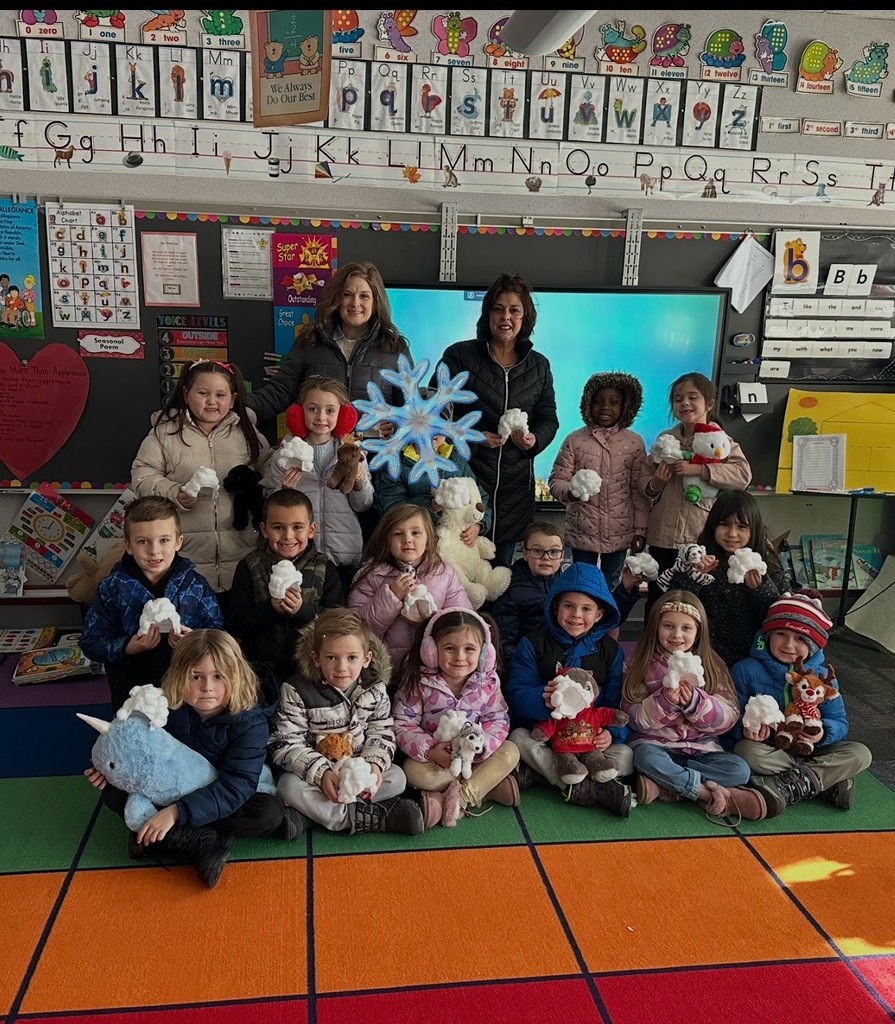 Students in a group photo wearing jackets holding Arctic stuff animals and igloo crafts with their teachers on the carpet in the classroom.