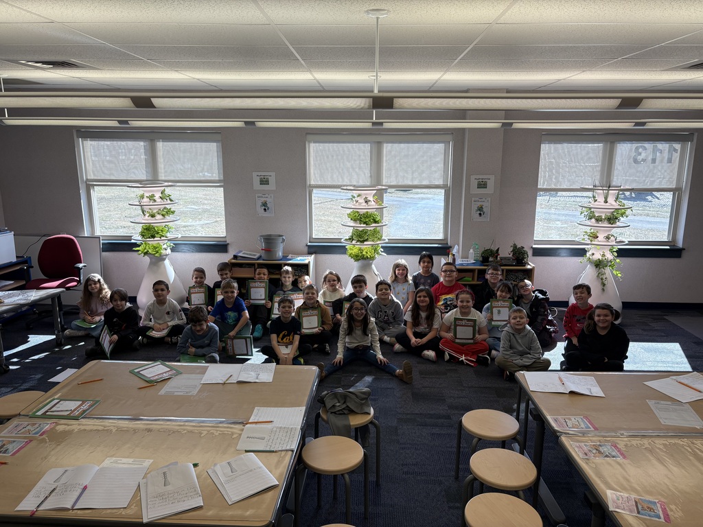 Students posing near hydroponic farm stand with lettuce growth and journals. 