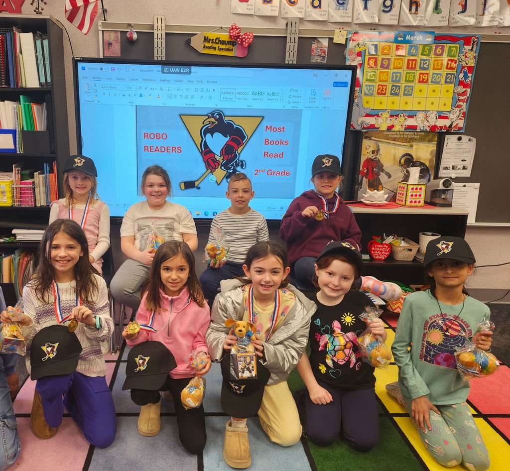 A group of students smilingv at the camera in front of a slide that says Robo Readers Most books read with a penguins hockey logo