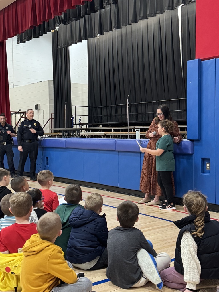A student presenting a thank you card to police officers in a gym with students seated in front of a stage 