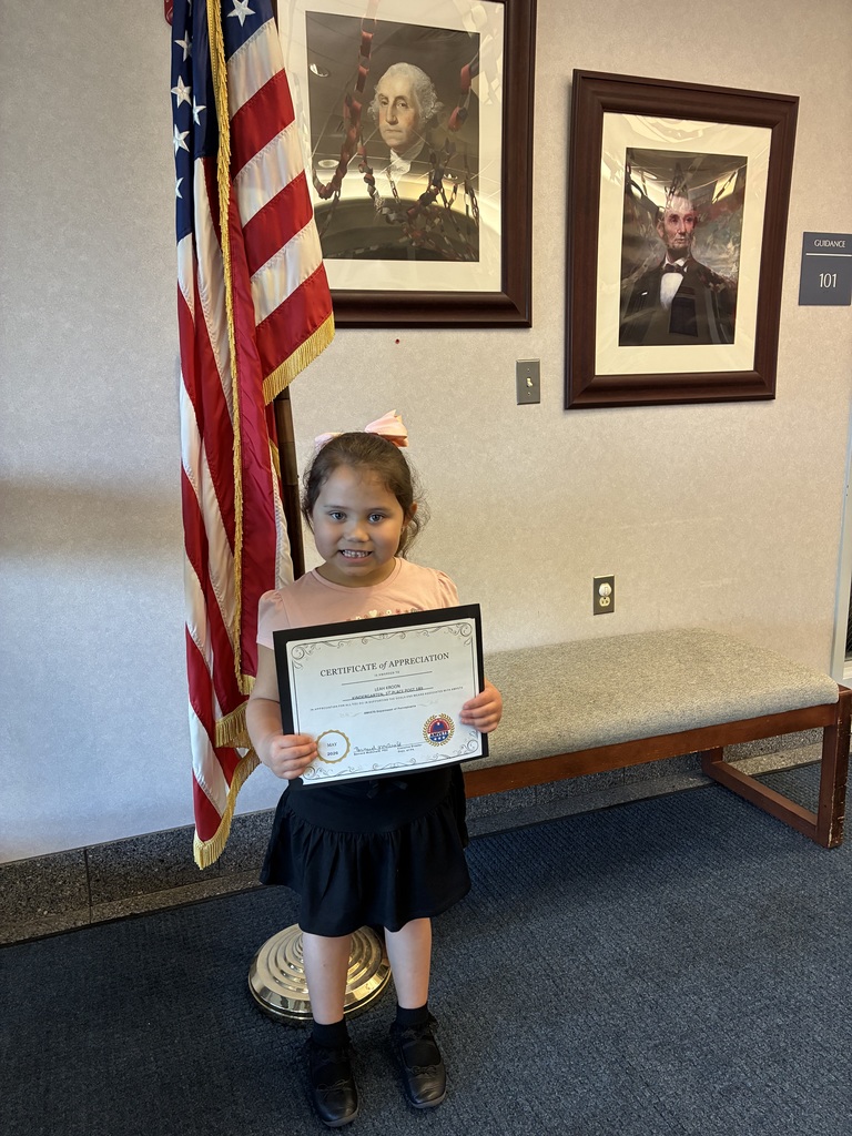 A student holding an amvets winner certificate in front of two president portraits and an american flag. 