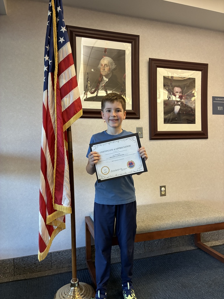 A student holding an amvets winner certificate in front of two president portraits and an american flag. 