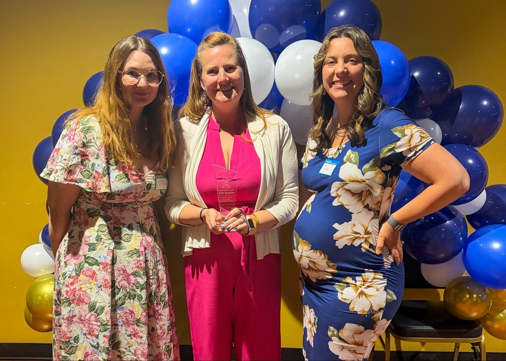 three woman stand in front of a blue, gold, and white balloon arch