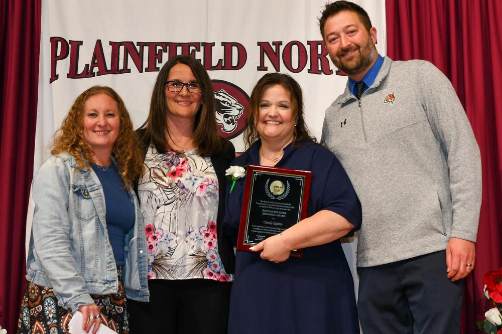 group of four teachers stand in front of Plainfield North High School backdrop. Woman in middle holds awards plaque