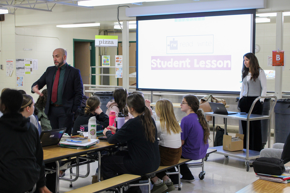 group of student seated listening to man speak while woman to rigtht stand by large screen