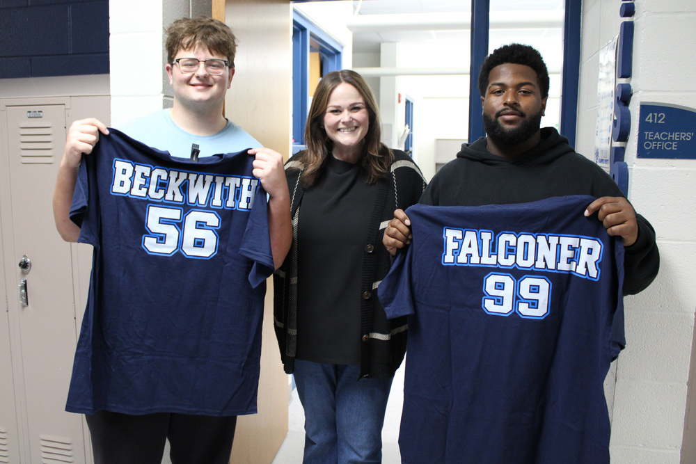 teacher stand between two Plainfield South High School football players holding shirts that say from left Beckwith 56, Falconer 99 on right