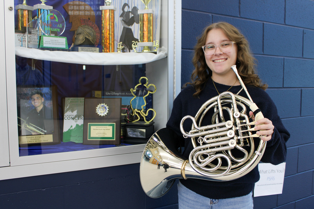 girl holds French horn standing near trophy case