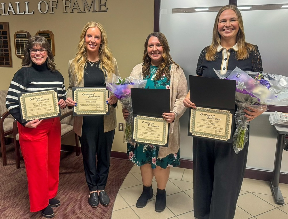 Four women stand in line holding certificates. Two hold flowers