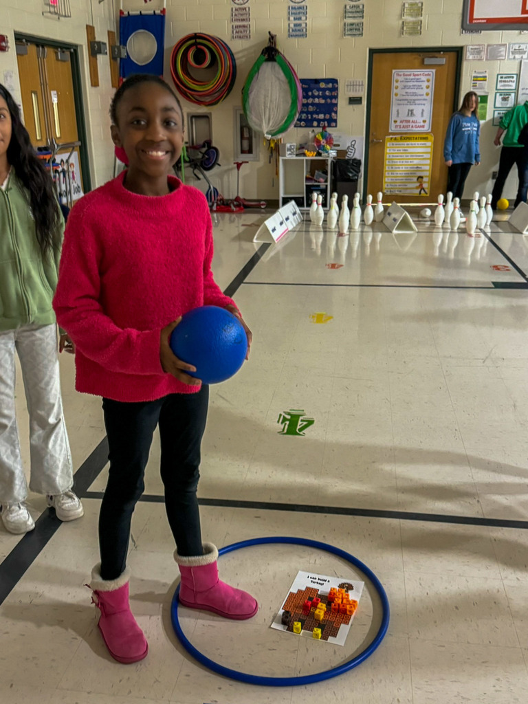 One student holding ball stands next to hula hoop with paper inside it