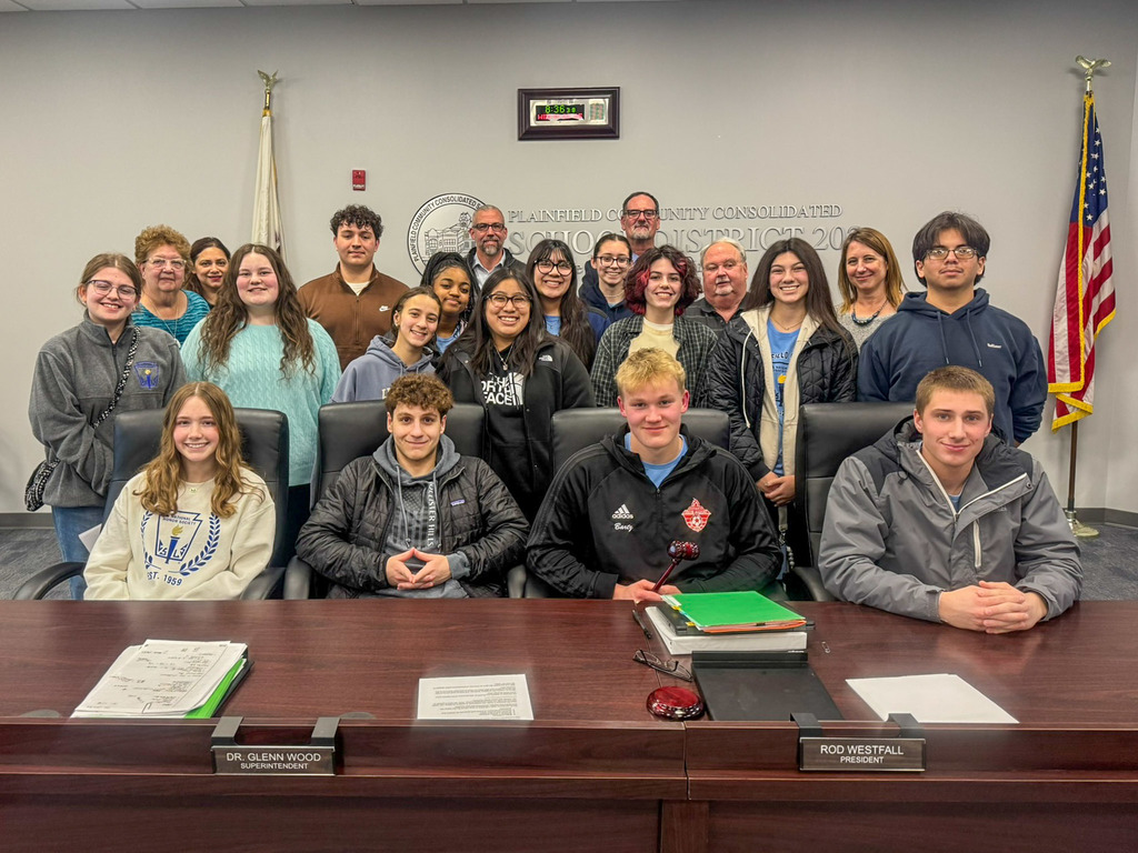 group of students seated at school board table. Adults stand in back of them