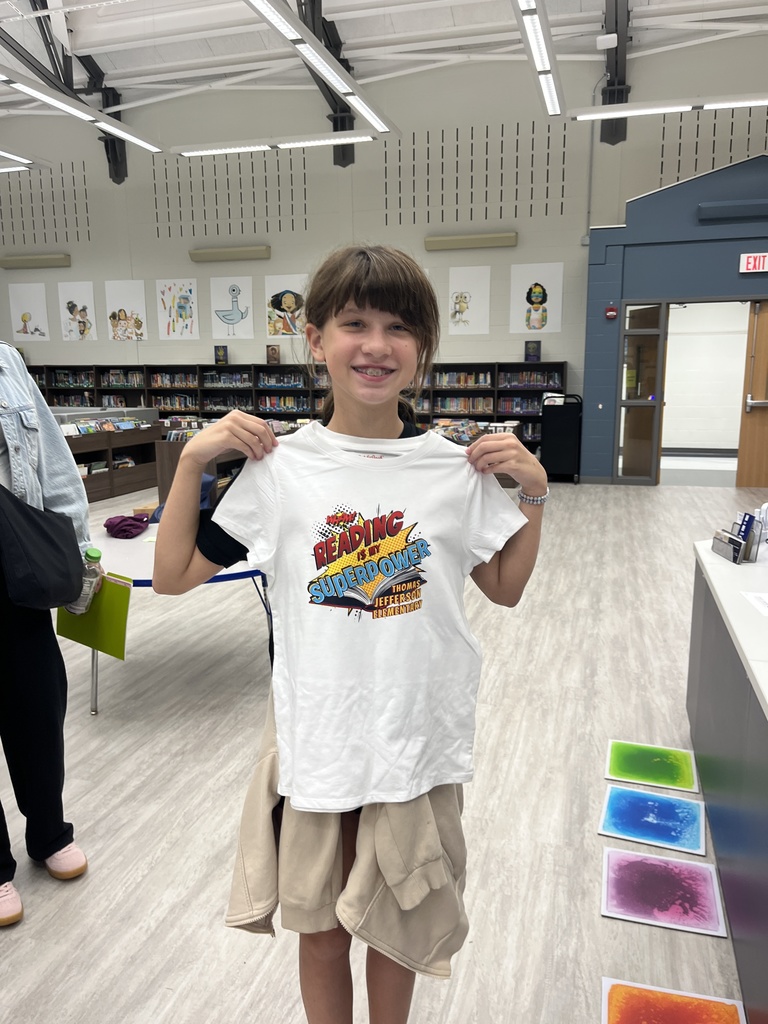 Student is standing in a library holding a T-shirt that says reading is my superpower.