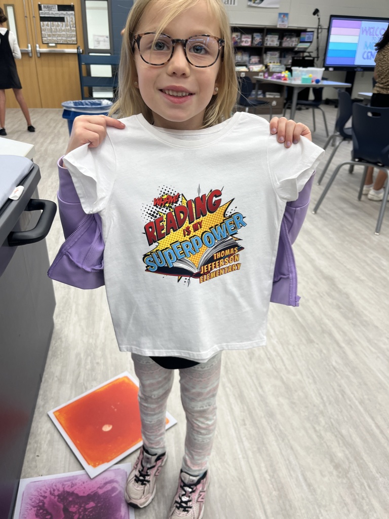 A student is holding a T-shirt that says reading is my superpower. 