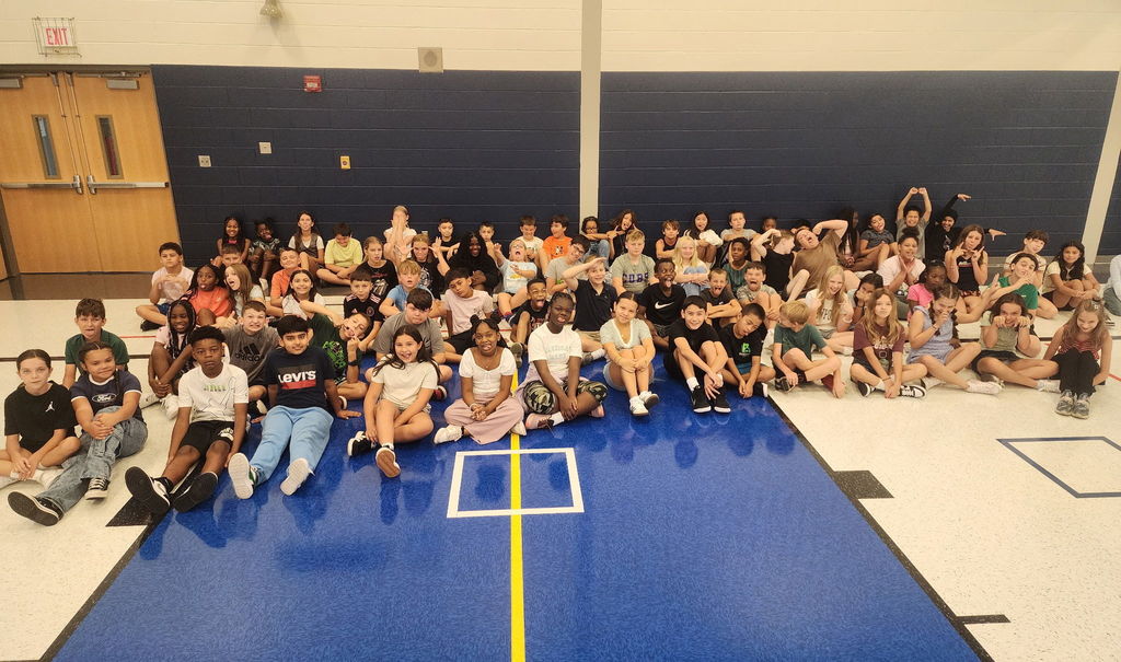 Many students are sitting on the floor of a gymnasium,