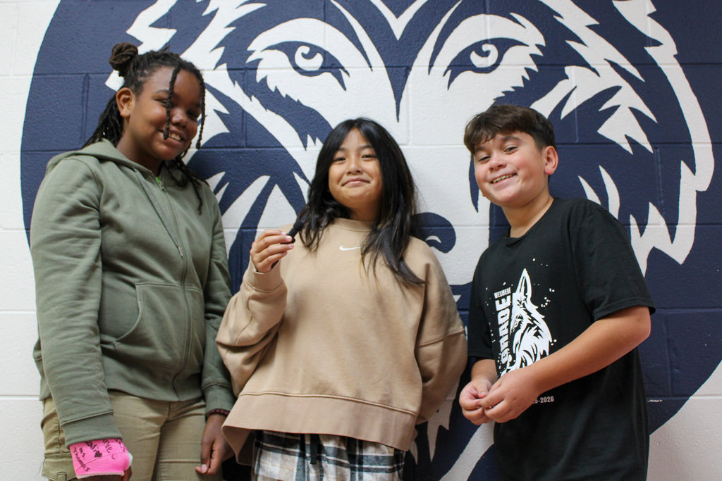 two female students and one male students stand in front of Wesmere Wolf logo