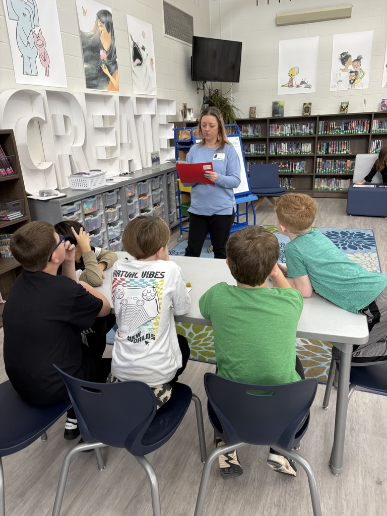 Students are sitting at a table in a library, and an adult is asking a question. 